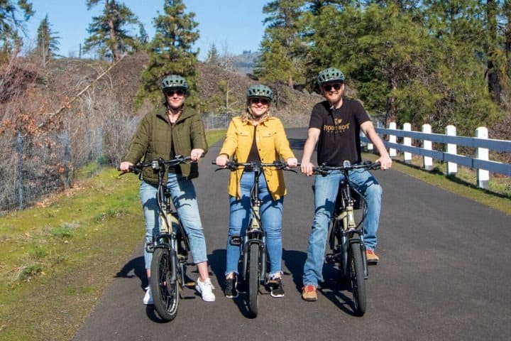 Three people wearing helmets ride e-bikes on a paved path surrounded by trees.