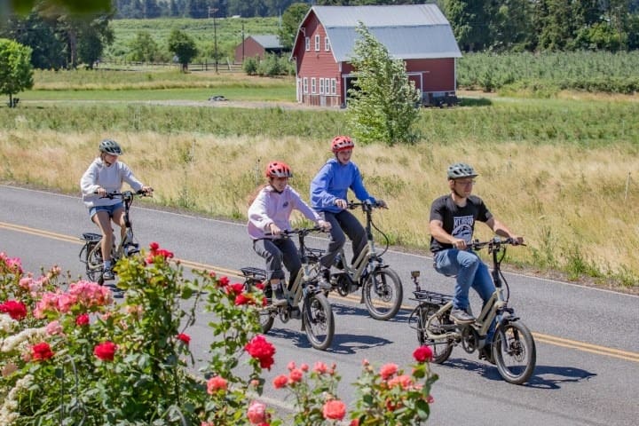 Four people ride electro bicycles on a rural road near a red barn and fields.