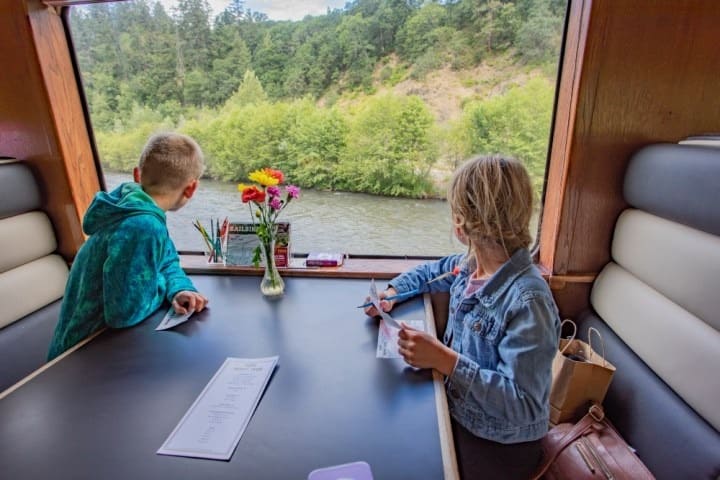 Two children sitting by a window on a train with a river and trees view, table with flowers and papers.