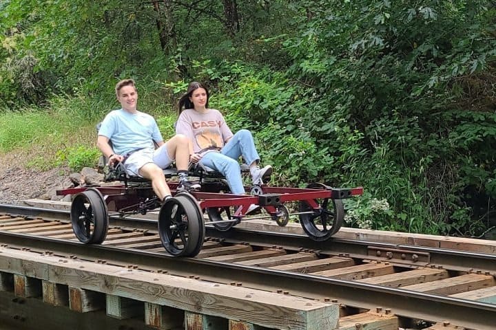 Two people riding a pedal-powered rail vehicle on train tracks in a forested area.