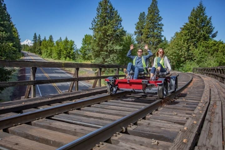 Two people ride a pedal cart on train tracks through a forested area.